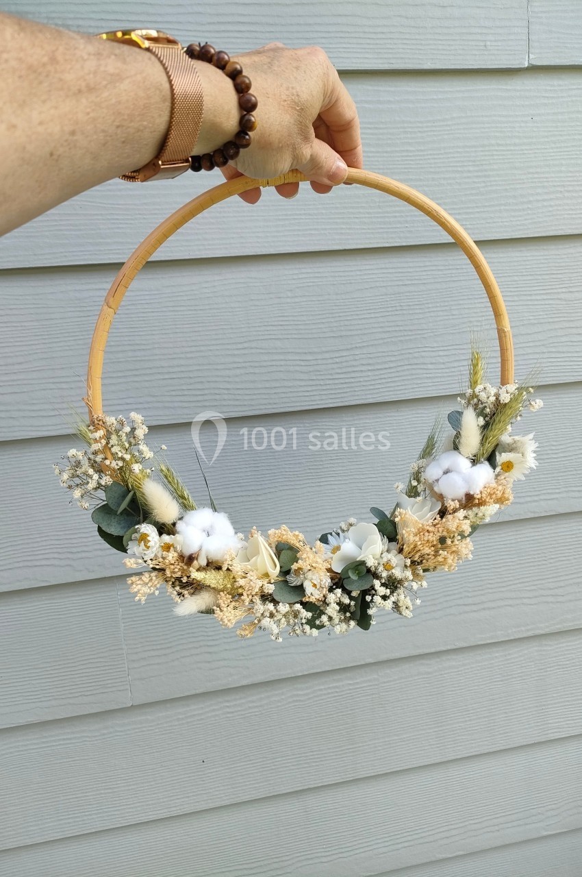 Couronne décorative en fleurs séchées et coton, tenue à la main devant un mur en bois gris.