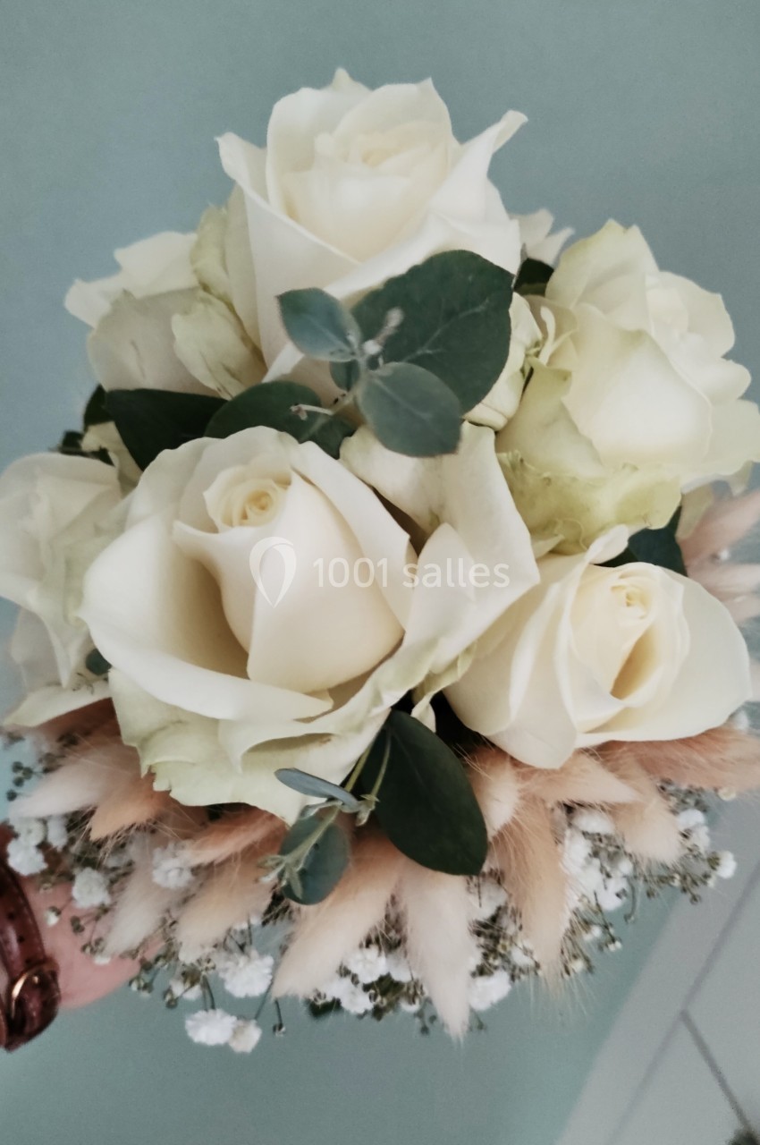 Bouquet de roses blanches entourées de feuillage vert, de gypsophile et de fleurs séchées beige, tenu à la main.