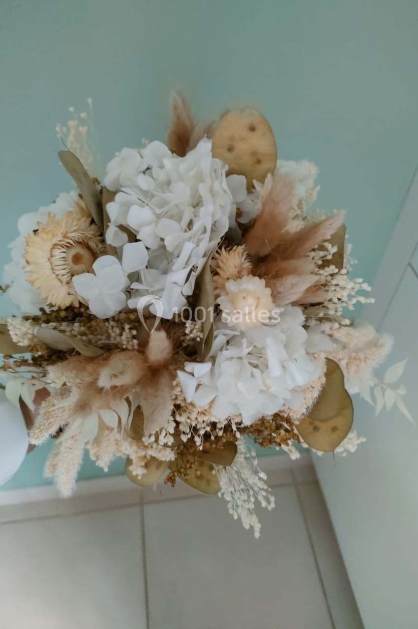 Bouquet de fleurs séchées et blanches, composé de diverses textures, tenu devant un mur bleu clair.