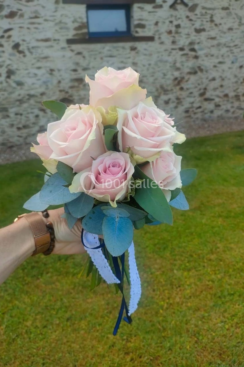 Bouquet de roses roses pâles avec feuillage vert, tenu par une main devant un mur en pierre et une pelouse.