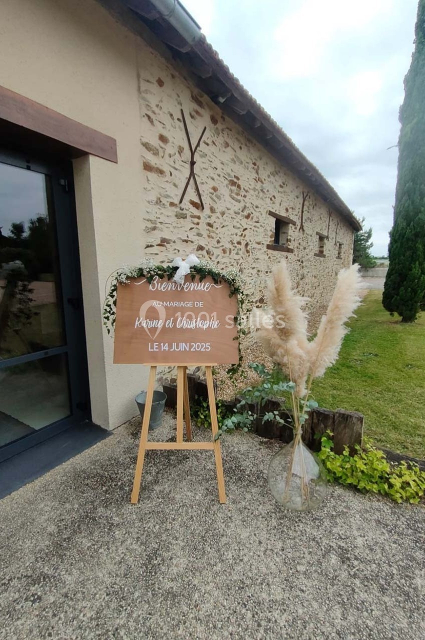Panneau de bienvenue en bois décoré de fleurs, posé sur un chevalet devant un bâtiment en pierre.