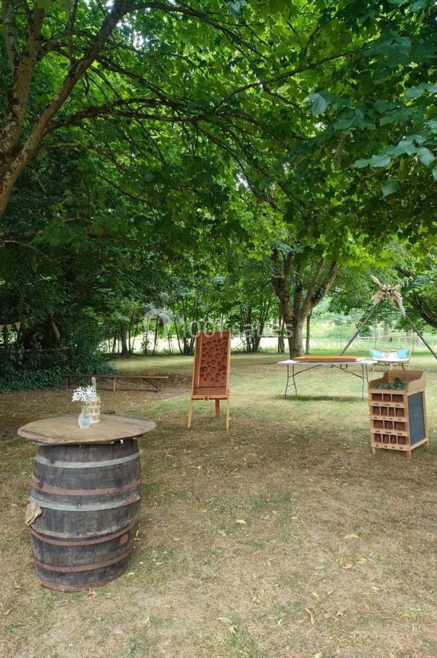 Espace extérieur avec tonneau en bois, présentoirs en bois et tables disposés sous des arbres dans un cadre naturel.