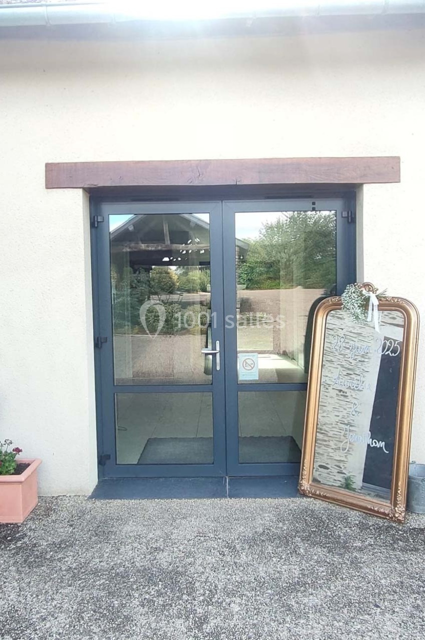 Porte vitrée double avec cadre gris, entourée de murs clairs, et miroir doré posé à droite affichant des inscriptions.