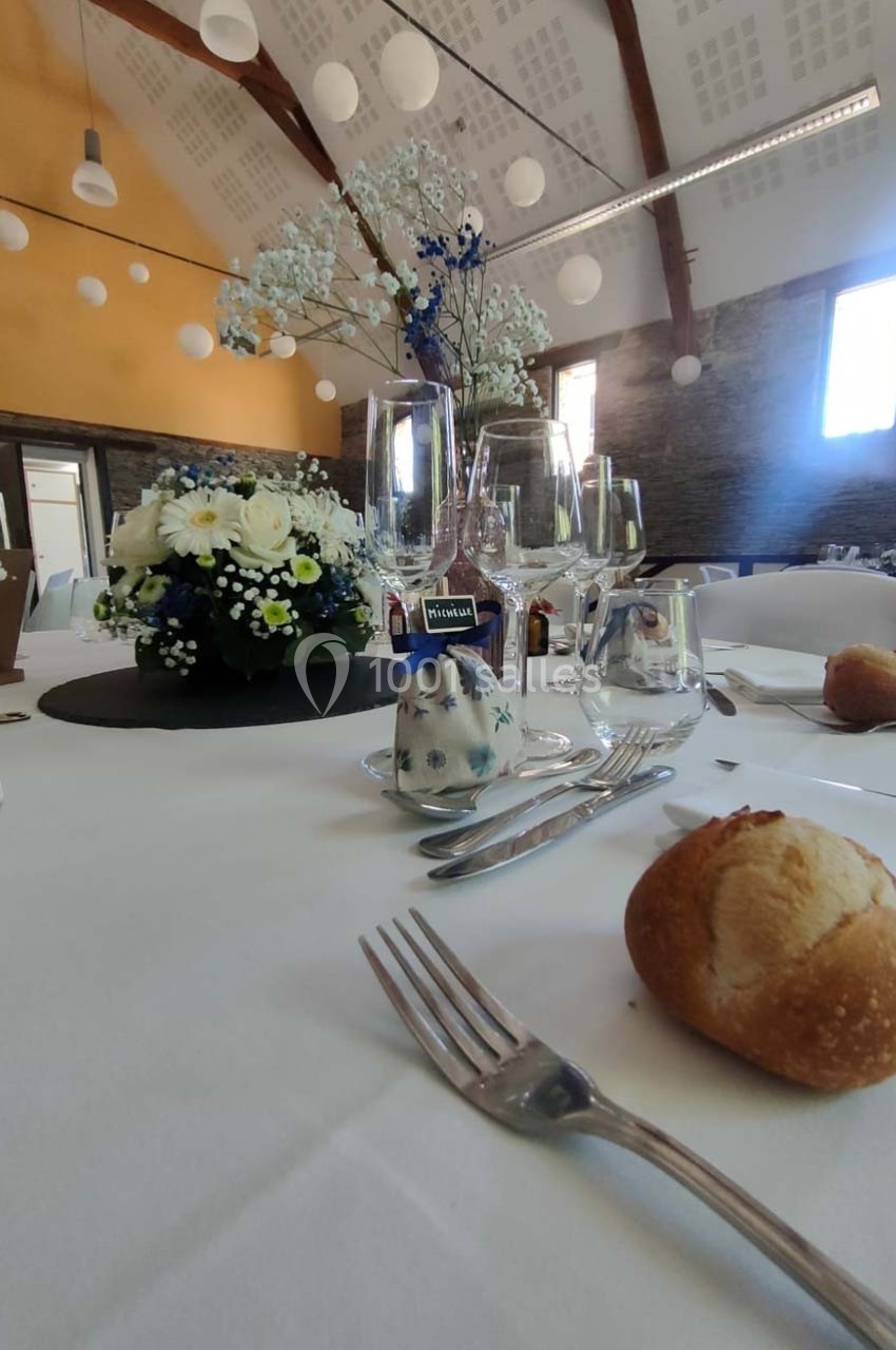 Table dressée avec nappes blanches, fleurs, verres, couverts et petits pains dans une salle lumineuse au plafond en poutres…
