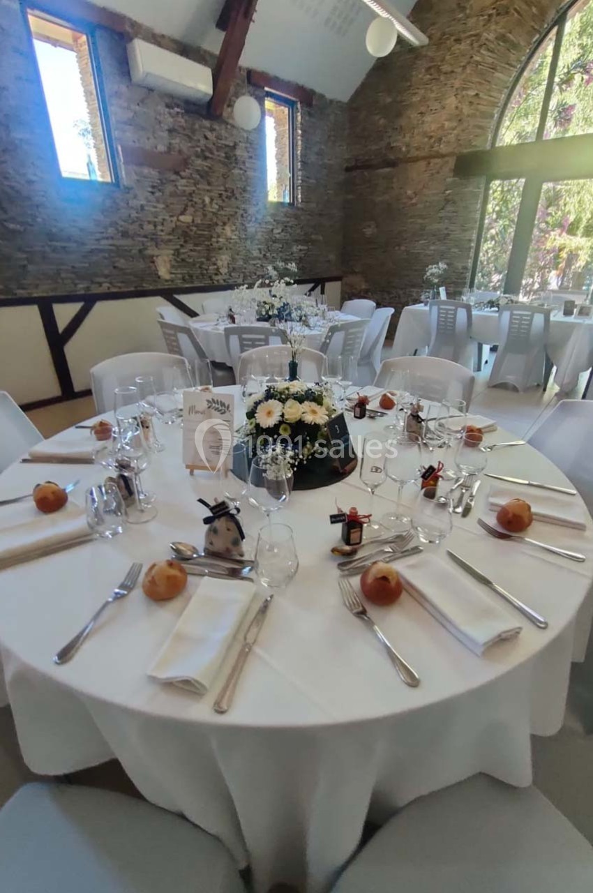 Salle de réception avec tables rondes dressées, nappes blanches, centres de table fleuris et lumière naturelle.