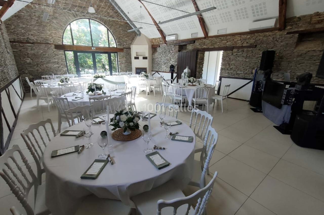 Salle de réception lumineuse avec tables rondes dressées, décorées de fleurs blanches et chaises blanches.
