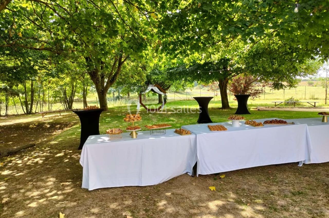Tables dressées avec des nappes blanches et des buffets de nourriture sous des arbres dans un espace extérieur ombragé.