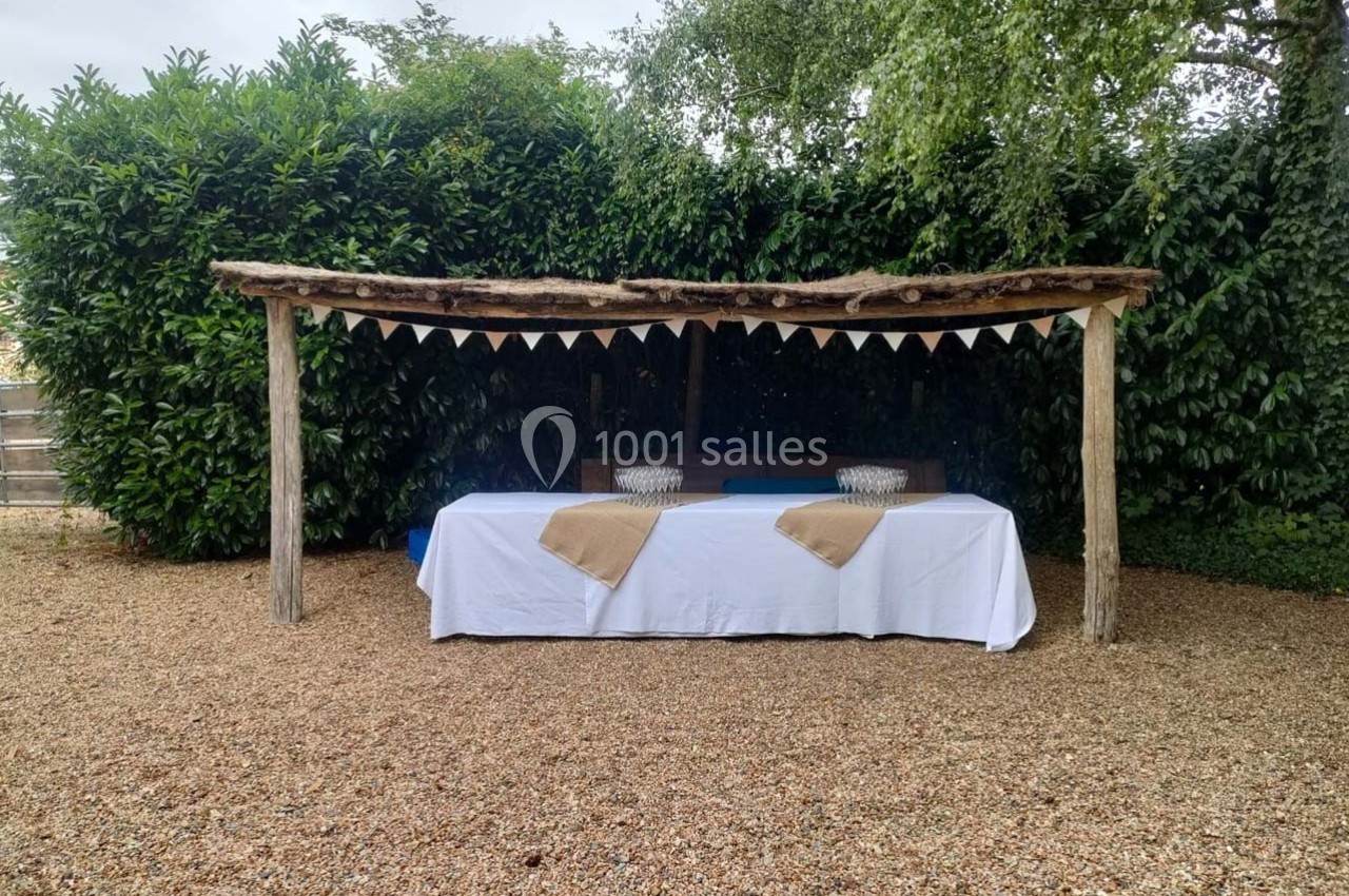 Table recouverte d'une nappe blanche sous une pergola en bois, décorée de fanions, sur un sol en gravier.