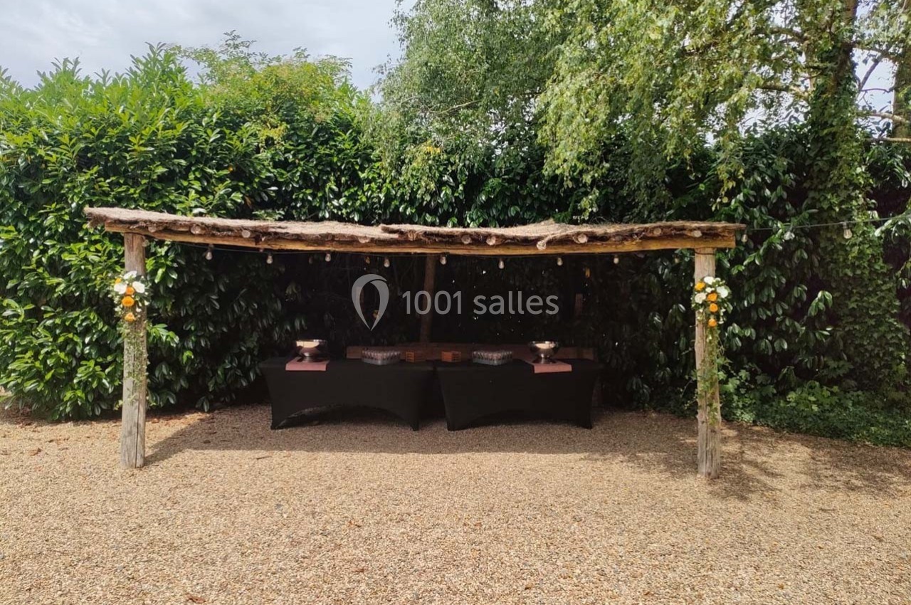 Abri en bois rustique avec tables noires dressées, entouré de végétation et décoré de fleurs.