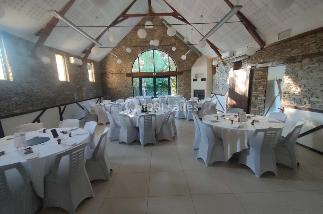 Salle de réception lumineuse avec tables rondes dressées et chaises blanches, murs en pierre et grande baie vitrée.