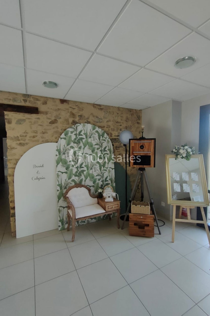 Coin photo décoré avec un fond végétal, un banc, une borne photo vintage et un tableau d'affichage.