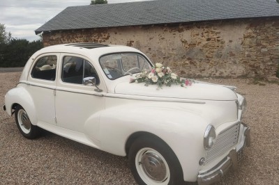 Voiture ancienne blanche décorée de fleurs, stationnée devant un bâtiment en pierre avec un toit en ardoise.
