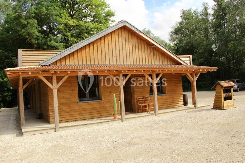 Bâtiment en bois avec une terrasse couverte, entouré d'arbres, sur un sol en gravier.
