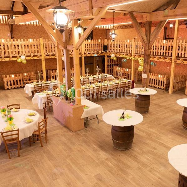 Salle de réception en bois avec tables rondes dressées et décorations festives, éclairée par des suspensions.