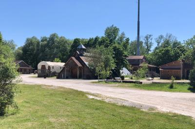 Bâtiments en bois avec une chapelle, une roulotte et des bottes de foin, entourés de verdure.
