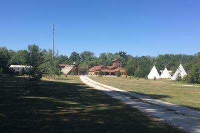 Bâtiments en bois avec une chapelle, une roulotte et des bottes de foin, entourés de verdure.