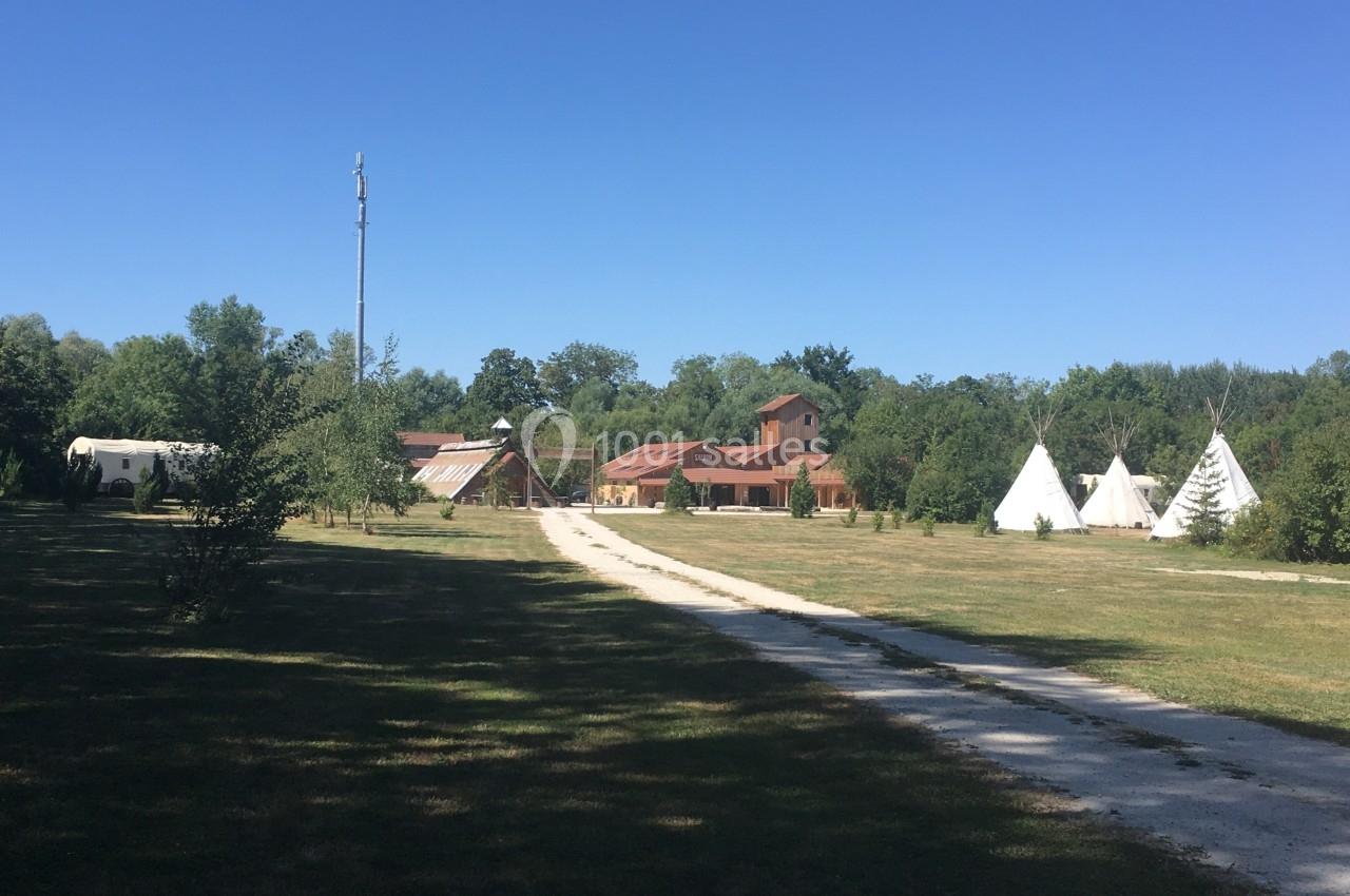 Chemin en gravier menant à des bâtiments en briques et des tipis blancs dans un environnement arboré.