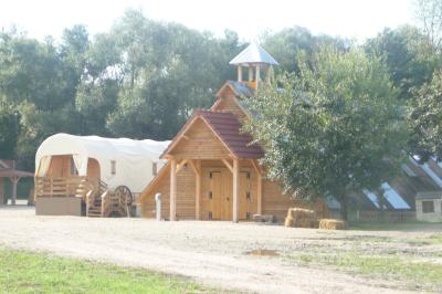 Bâtiments en bois avec une chapelle, une roulotte et des bottes de foin, entourés de verdure.
