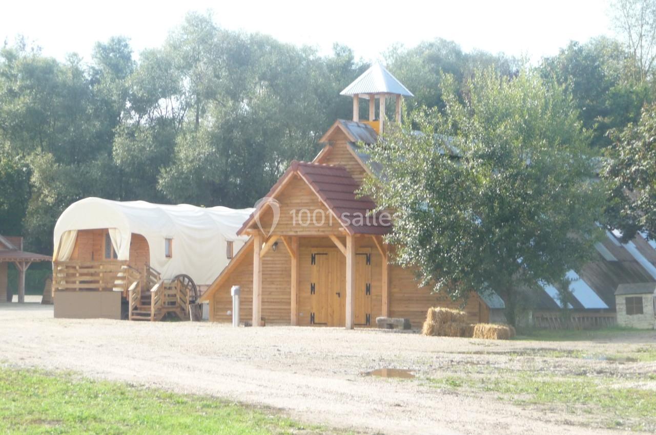 Bâtiments en bois avec une chapelle, une roulotte et des bottes de foin, entourés de verdure.