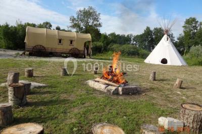 Bâtiments en bois avec une chapelle, une roulotte et des bottes de foin, entourés de verdure.