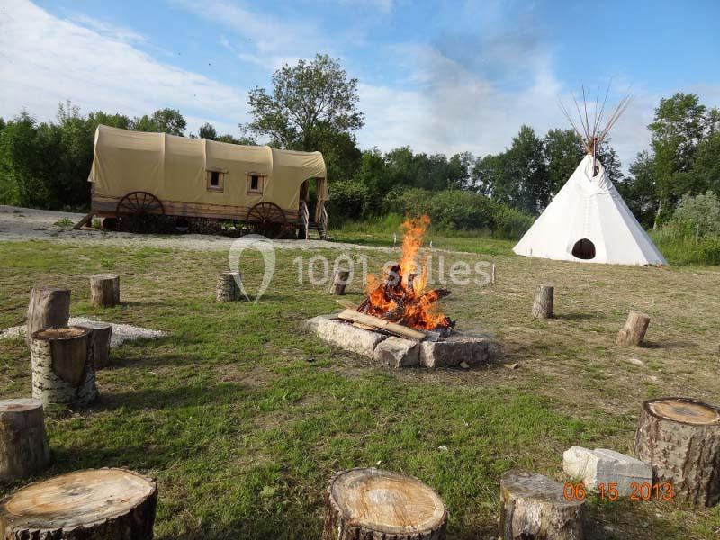 Feu de camp entouré de rondins, avec une roulotte et un tipi en arrière-plan dans un cadre naturel.