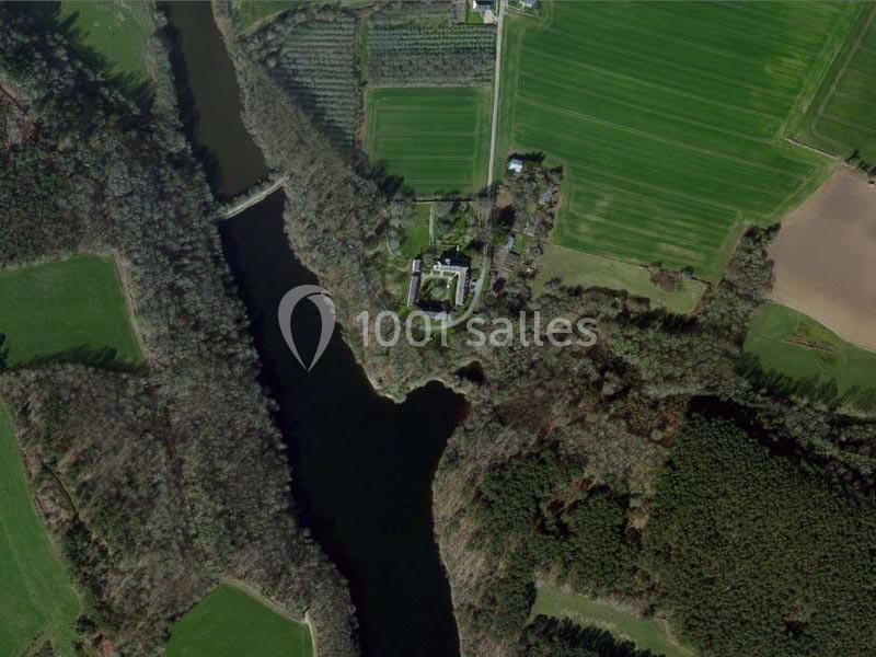 Vue aérienne d'un paysage rural avec un bâtiment entouré d'arbres, près d'une rivière et de champs cultivés.