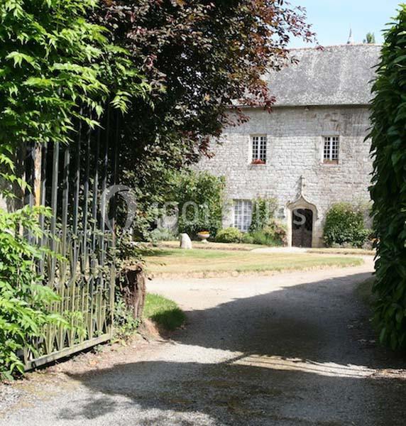 Allée bordée de verdure menant à une maison en pierre avec toit en ardoise et fenêtres à petits carreaux.