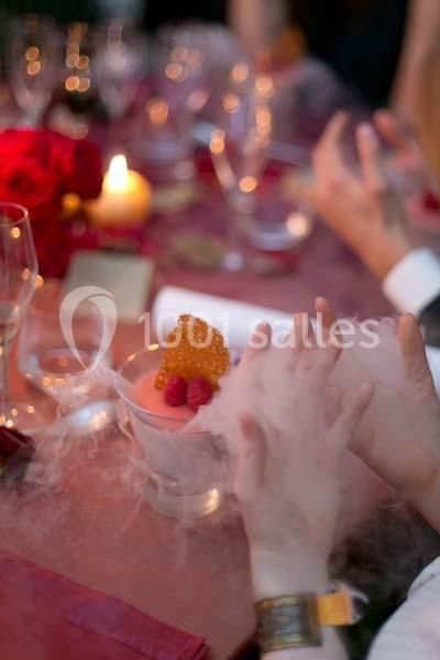 Une main effleure un dessert entouré de fumée, décoré de framboises et d'une tuile croustillante, sur une table élégante.