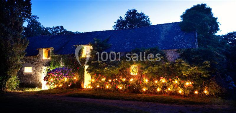 Façade d'une maison en pierre éclairée par des guirlandes lumineuses et entourée de végétation au crépuscule.
