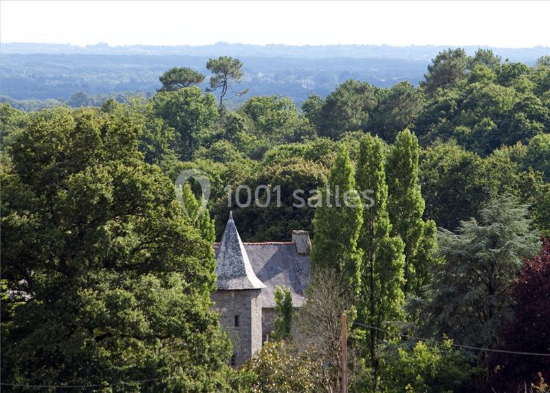 Vue d'un paysage boisé avec un bâtiment en pierre et un toit en ardoise partiellement visible au premier plan.
