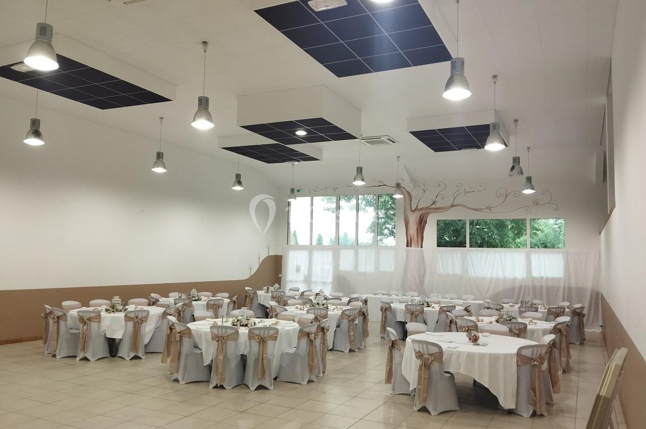 Salle de réception lumineuse avec tables rondes dressées, chaises décorées de nœuds beiges et fresque d'arbre sur un mur.