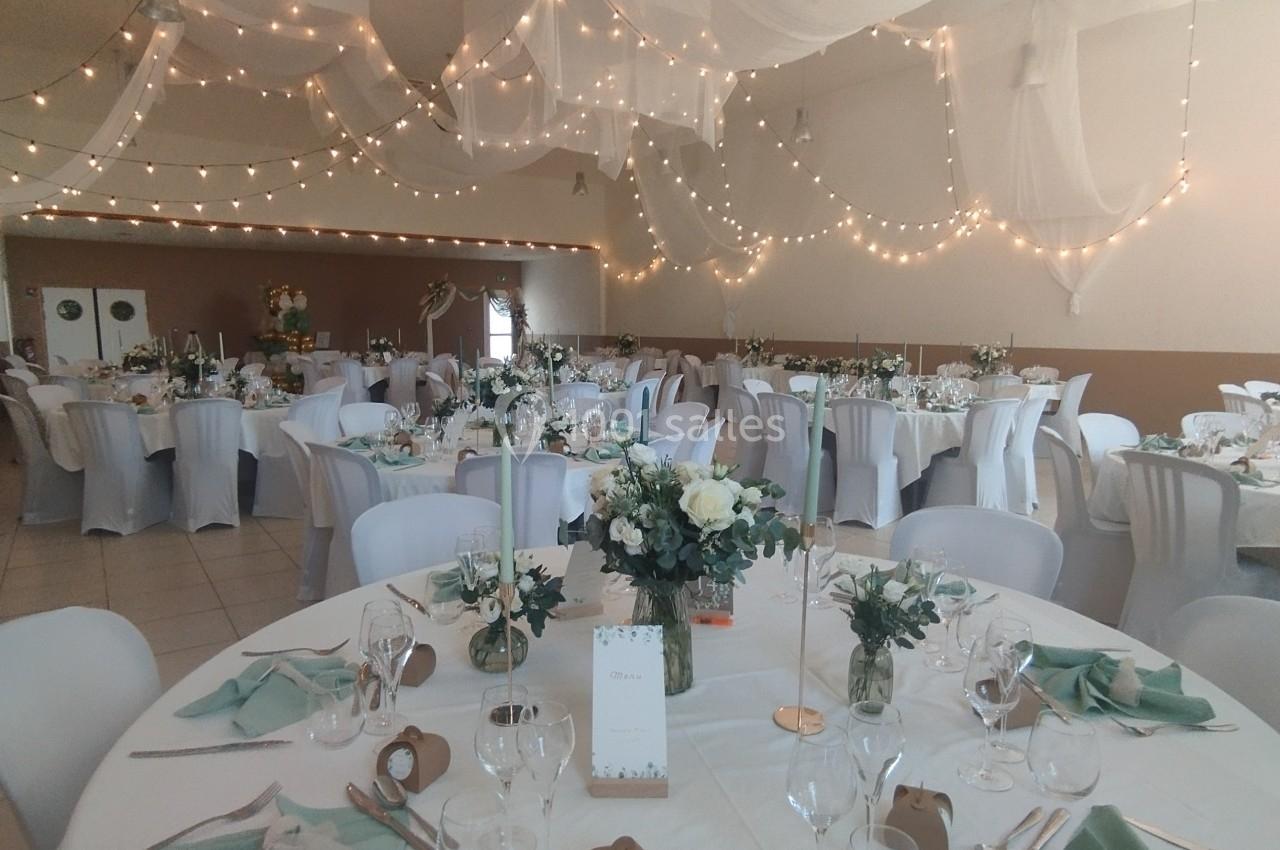 Salle de réception décorée avec des tables rondes, nappes blanches, serviettes vertes et guirlandes lumineuses suspendues.