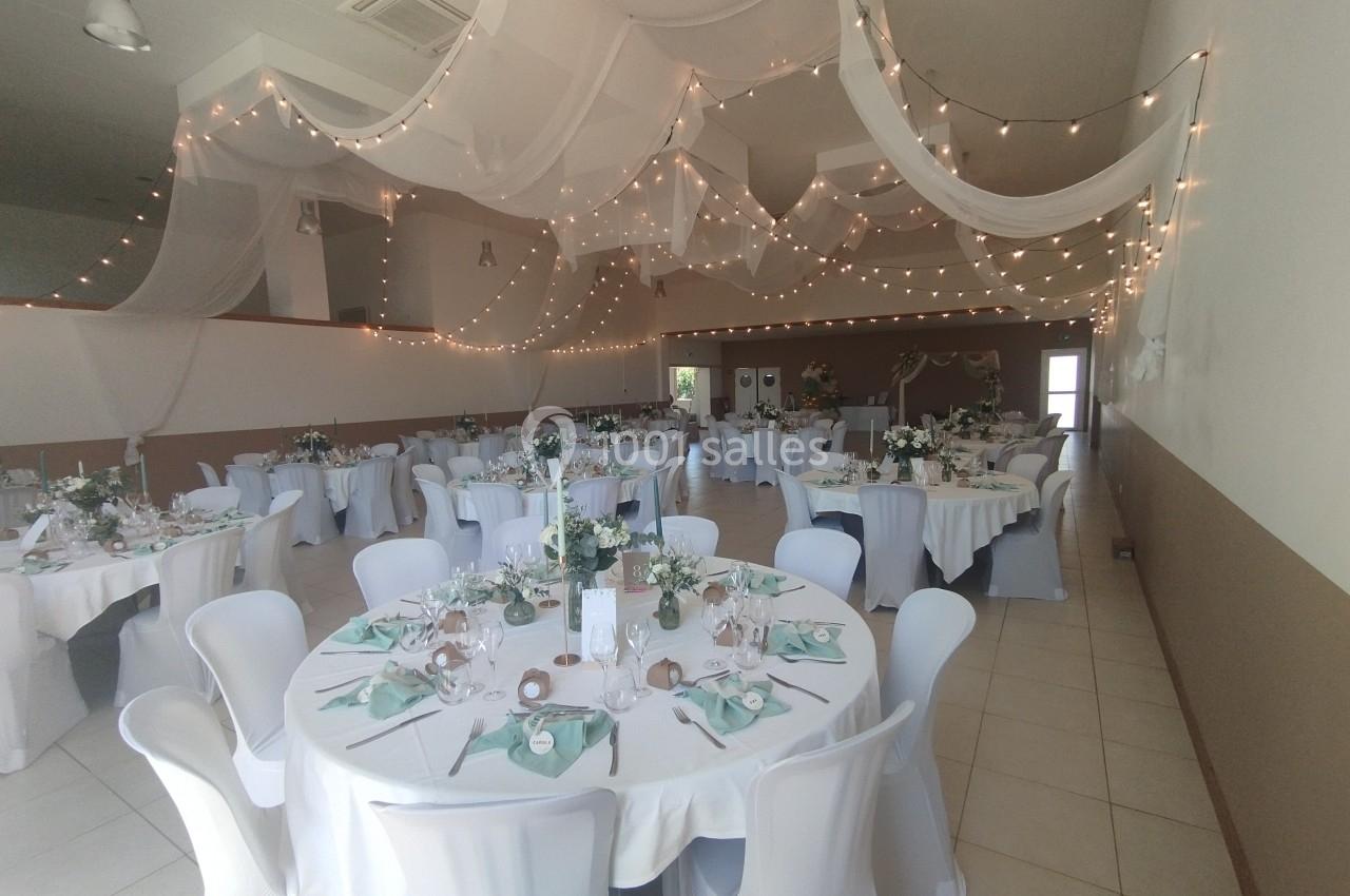 Salle de réception décorée avec des guirlandes lumineuses, des nappes blanches et des tables dressées pour un événement.