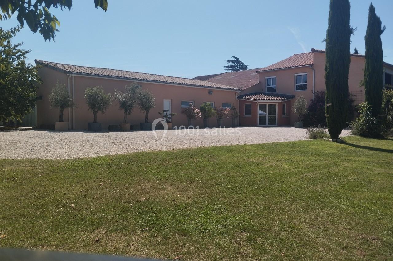 Grande maison rose avec toit en tuiles, entourée d'arbres, pelouse et allée en gravier sous un ciel dégagé.