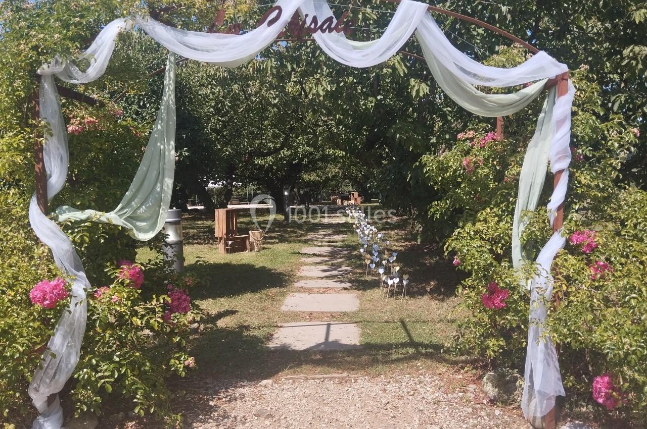 Allée de jardin bordée de fleurs et d'arbres, avec une arche décorée de voilages blancs au premier plan.