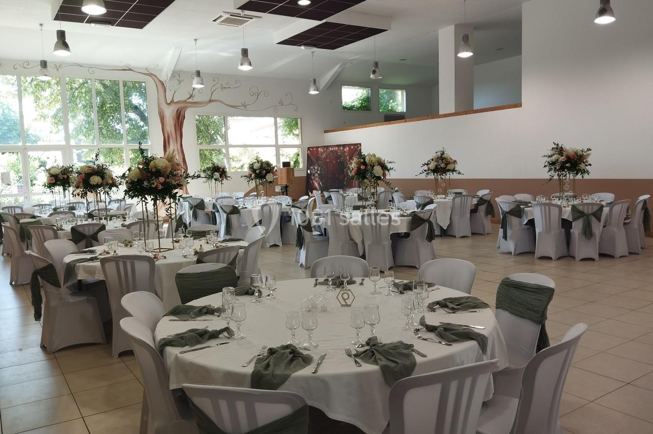 Salle de réception décorée avec des tables rondes, nappes blanches, chaises ornées de nœuds verts et centres de table…