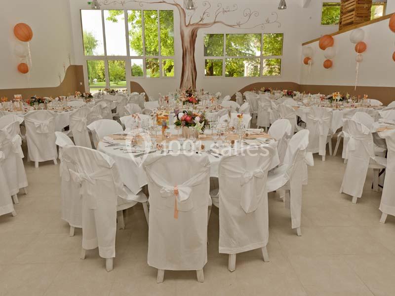 Salle de réception décorée avec des tables rondes, nappes blanches, chaises habillées et ballons suspendus.