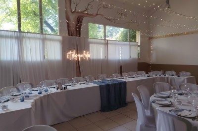 Salle décorée pour un événement, avec tables dressées, nappes noires, centres de table à plumes blanches et ballons dorés.