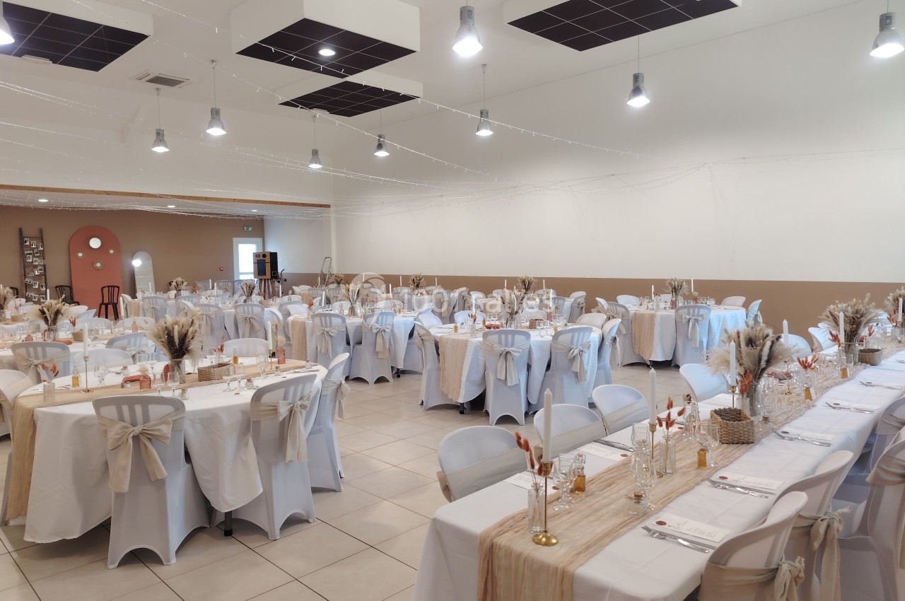 Salle de réception décorée pour un événement, avec tables dressées, nappes blanches et ornements beige et dorés.
