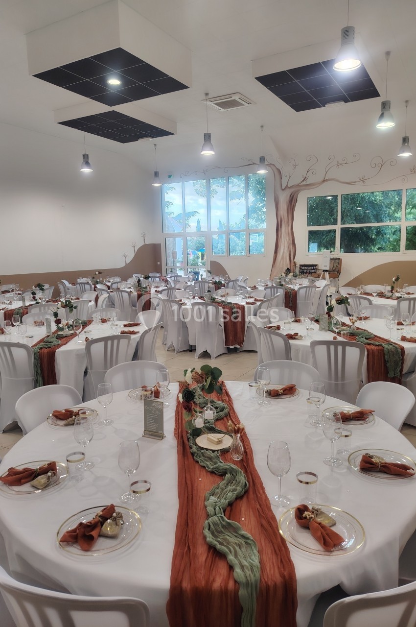 Salle de réception décorée avec des tables rondes, nappes blanches et chemins de table orange et verts.