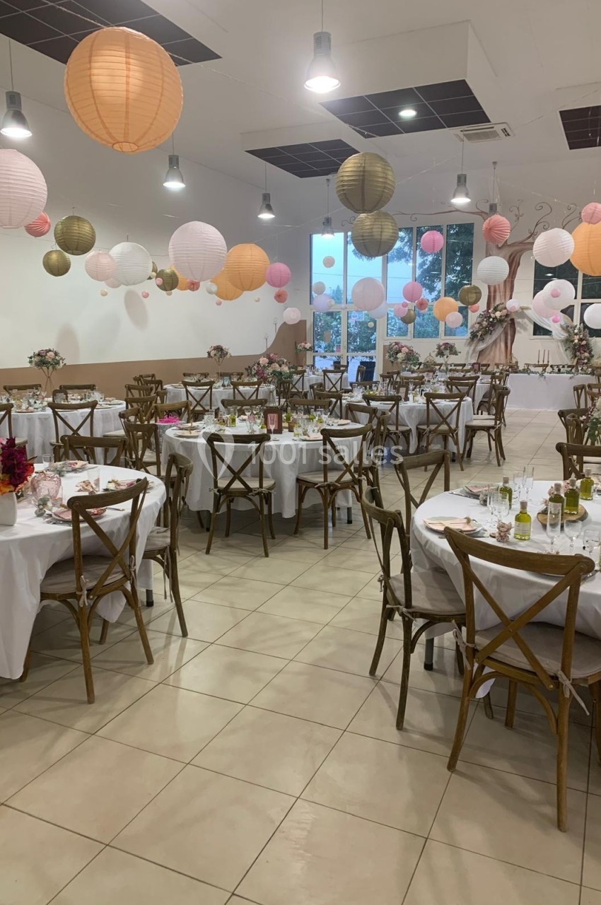 Salle décorée pour une réception avec tables rondes dressées, chaises en bois et lanternes suspendues colorées.