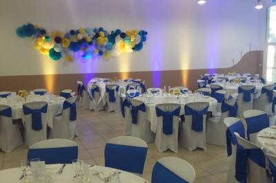 Salle décorée pour un événement, avec tables dressées, nappes noires, centres de table à plumes blanches et ballons dorés.