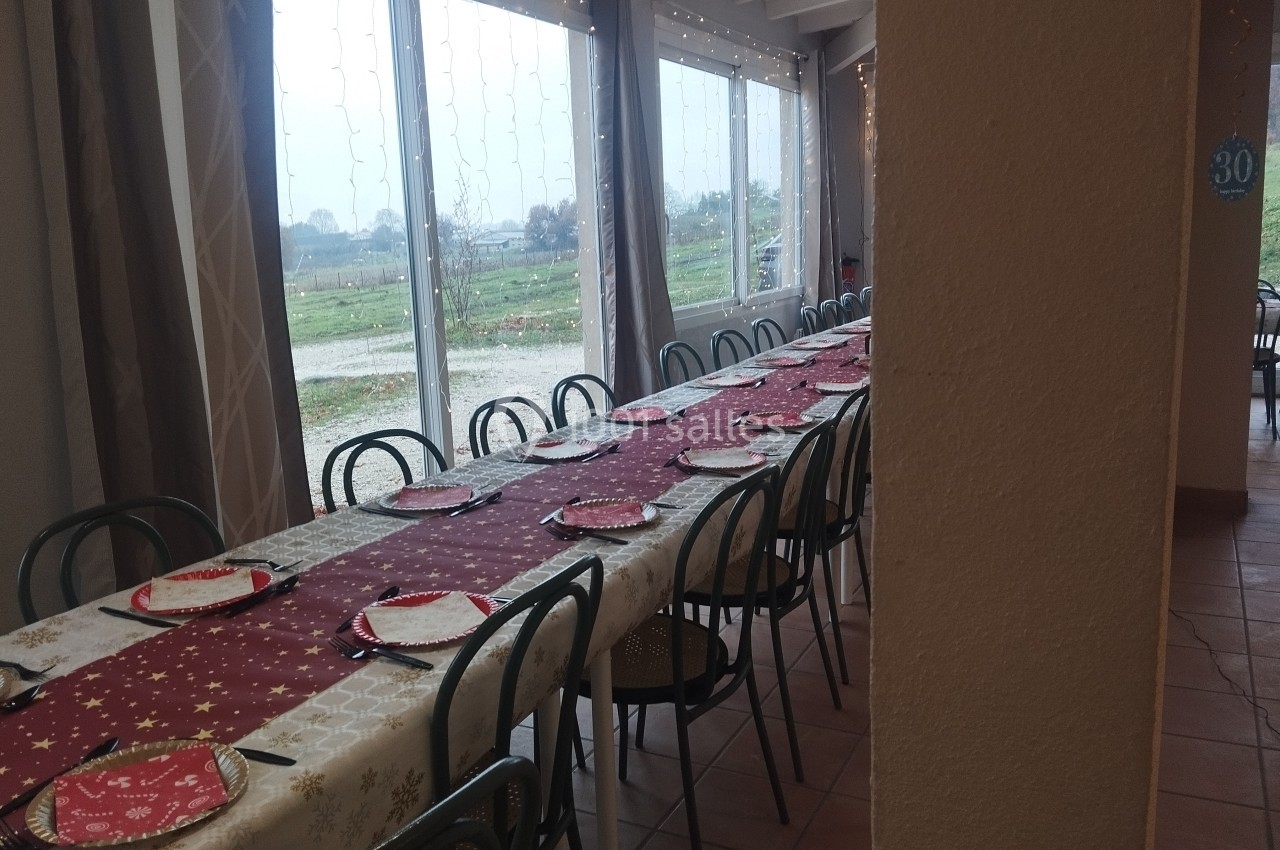 Grande table rectangulaire dressée avec nappes festives, assiettes et chaises, près de baies vitrées donnant sur un paysage…