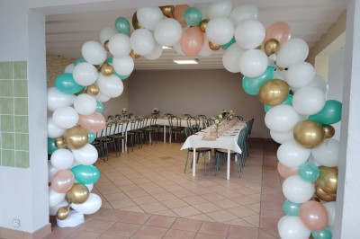 Salle décorée pour un événement, avec tables dressées, nappes noires, centres de table à plumes blanches et ballons dorés.