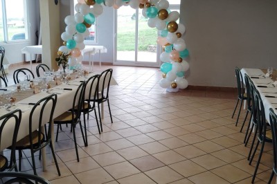 Salle décorée pour un événement, avec tables dressées, nappes noires, centres de table à plumes blanches et ballons dorés.
