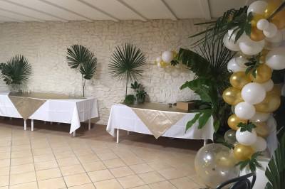 Salle décorée pour un événement, avec tables dressées, nappes noires, centres de table à plumes blanches et ballons dorés.