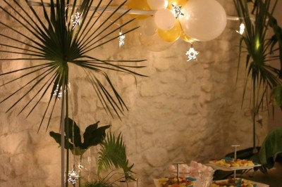 Salle décorée pour un événement, avec tables dressées, nappes noires, centres de table à plumes blanches et ballons dorés.