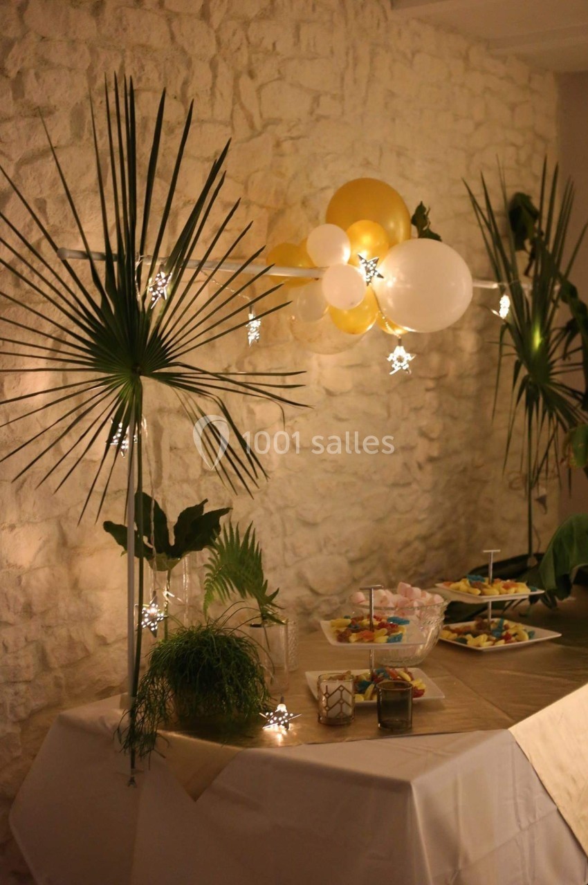 Table décorée avec des plantes, guirlandes lumineuses, ballons blancs et dorés, et plats disposés pour une réception.
