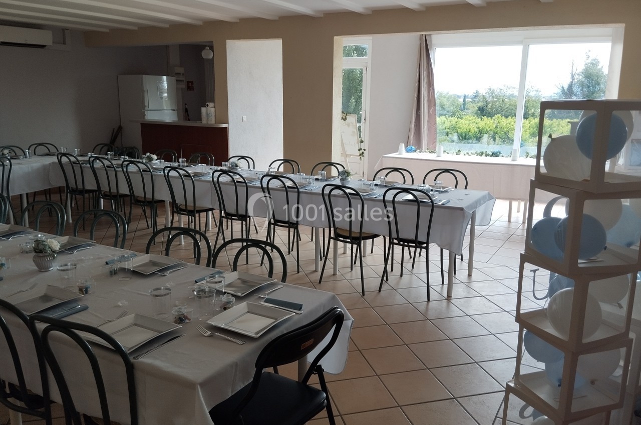 Salle lumineuse aménagée avec de longues tables dressées pour un repas, donnant sur un paysage extérieur verdoyant.