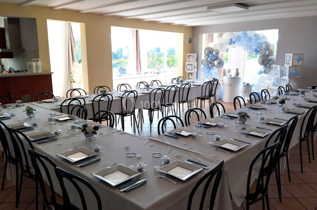Salle décorée pour un événement avec tables dressées en U, nappes blanches et ballons en arrière-plan.